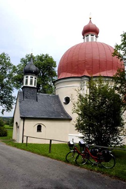 Eine Kapelle mit einem kleinen und einem großen Zwiebelturm auf der Strecke der Münchner Seenrunde - am Geländer davor sind zwei Fahrräder abgestellt.