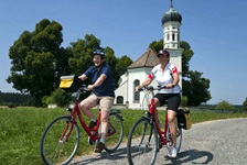 Zwei Radler fahren auf einem befestigten Radweg an einer Kapelle auf der Strecke der Münchner Seenrunde bzw. bayerischen Seenrunde