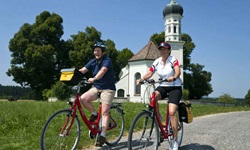 Zwei Radler fahren auf einem befestigten Radweg an einer Kapelle auf der Strecke der Münchner Seenrunde bzw. bayerischen Seenrunde