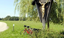Ein Feldkreuz mit Blumenschmuck auf einer Wiese, dahinter eine Bank mit zwei abgestellten Fahrrädern auf der Strecke der Münchner Seenrunde und der bayerischen Seenrunde