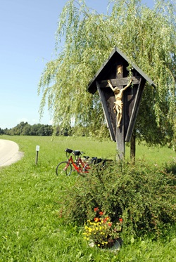 Ein Feldkreuz mit Blumenschmuck auf einer Wiese, dahinter eine Bank mit zwei abgestellten Fahrrädern auf der Strecke der Münchner Seenrunde und der bayerischen Seenrunde