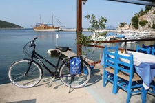 Ein Fahrrad steht an der Promenade zum Meer an einem Restaurant. Im Hintergrund ist das angelegte Schiff Panagiota zu sehen