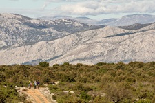 Mountainbiker fahren auf einem Schotterweg durch die Landschaft eines Nationalparks in Dalmatien hinab - hinter ihnen sind die Kulissen der gigantischen Berge Kroatiens zu sehen