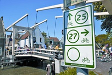 Eine Gruppe Radler fährt über eine Zugbrücke in Holland, im Vordergrund auf der rechten Seite ist ein Fahrradschild mit den Routen Nr. 25 und 27 und der Aufschrift NOORD-HOLLAND zu sehen