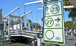 Eine Gruppe Radler fährt über eine Zugbrücke in Holland, im Vordergrund auf der rechten Seite ist ein Fahrradschild mit den Routen Nr. 25 und 27 und der Aufschrift NOORD-HOLLAND zu sehen