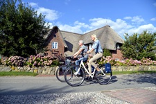 Zwei Radler fahren an einem reetgedeckten Haus mit herrlichem Blumenschmuck vorbei.