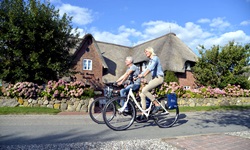 Zwei Radler fahren an einem reetgedeckten Haus mit herrlichem Blumenschmuck vorbei.