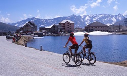 Zwei Radlerinnen fahren auf der Passhöhe des St. Gotthard am Seeufer entlang auf das Gotthard-Hospiz und das St-Gotthard-Museum zu. Im Hintergrund erheben sich schneebedeckte Alpengipfel.