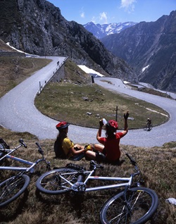 Zwei Radlerinnen haben ihre Abfahrt vom St. Gotthard für eine kurze Trinkpause unterbrochen und sitzen am Rand einer Serpentinenkurve.