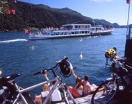 Radler sitzen am Ufer eines Sees im Tessin auf einem Steg und winken den vorbeifahrenden Schiffen zu.