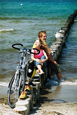 Zwei Personen sitzen auf Holzpflöcken an der Ostsee - neben ihnen steht ein Fahrrad