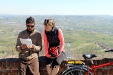 Zwei Radfahrer machen eine Pause an einer Mauer und blicken auf ihre Unterlagen - im Hintergrund ist die Landschaft mit Orten des Piemonts zu sehen