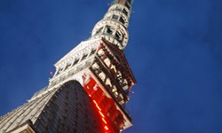 Detailbild auf eines der Wahrzeichen Turins - die in der Dämmerung rot beleuchtete Mole Antonelliana mit ihrer markanten Turmspitze, welche mit einem fünfzackigen Stern gekrönt ist.