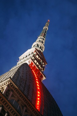 Detailbild auf eines der Wahrzeichen Turins - die in der Dämmerung rot beleuchtete Mole Antonelliana mit ihrer markanten Turmspitze, welche mit einem fünfzackigen Stern gekrönt ist.