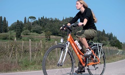 Eine Radfahrerin fährt auf einer Straße an einem mit Zypressen bewachsenen Hügel in der Toskana entlang.