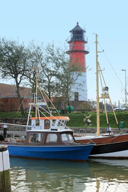 Zwei Boote sind an einem kleinen Hafen am Nordseeküsten-Radweg angelegt, im Hintergrund ist ein rot-weißer Leuchtturm zu sehen