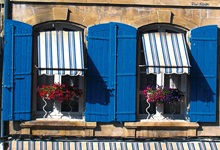 Häuserfassade mit zwei blumengeschückten Fenstern und blauen Holzläden sowie einem weiß-blau gestreiften Sonneschutz