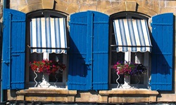 Häuserfassade mit zwei blumengeschückten Fenstern und blauen Holzläden sowie einem weiß-blau gestreiften Sonneschutz