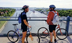 Zwei Radfahrer stehen auf einer Brücke bei der Radreise Ostseeküste und Holsteinische Schweiz und sie blicken auf den Kanal mit Schiffen