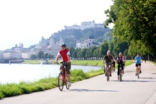 Eine Gruppe Radfahrer auf einem asphaltierten Radweg bei Salzburg. Im Hintergrund ist die Skyline der Mozartstadt zu erkennen.