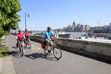 Drei Radler fahren am Donauufer von Budapest entlang - auf der gegenüberliegenden Flussseite ist das Parlamentsgebäude zu erkennen.