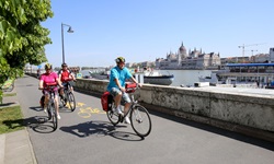 Drei Radler fahren am Donauufer von Budapest entlang - auf der gegenüberliegenden Flussseite ist das Parlamentsgebäude zu erkennen.