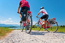 Zwei Radler fahren auf einem weiß gekiesten Radweg im bayerischen Voralpenland auf einen See zu.