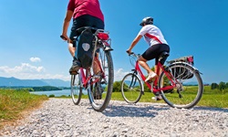 Zwei Radler fahren auf einem weiß gekiesten Radweg im bayerischen Voralpenland auf einen See zu.