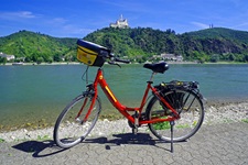 Ein rotes Fahrrad mit Lenkertasche steht am Ufer des Rheins mit Blick nach Goarshausen und seiner Höhenburg