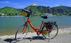 Ein rotes Fahrrad mit Lenkertasche steht am Ufer des Rheins mit Blick nach Goarshausen und seiner Höhenburg