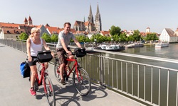Zwei Radfahrer radeln über eine Brücke in Regensburg über die Donau - im Hintergrund ist der Dom St. Peter mit seinen zwei Türmen zu sehen