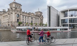 Drei Radfahrer stehen an der Spree, auf der ein Schiff fährt, und blicken auf den Reichstag