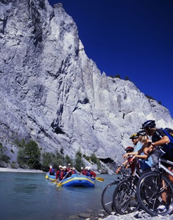 Eine Gruppe Radler hat am Schweizer Rheinufer vor einer majestätischen Bergkulisse angehalten und beobachtet, wie zwei Schlauchboote den Rhein überqueren.