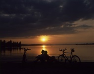 Zwei Radfahrer sitzen Rücken an Rücken und genießen einen wunderschönen Sonnenuntergang über dem Bodensee.