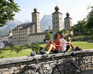 Zwei Radler - ein Mann und eine Frau - sitzen Rücken an Rücken auf einer Mauer vor dem bekannten Stockalperschloss von Brig mit seinen markanten drei Türmen.