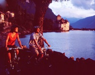 Ein Mann und eine Frau radeln am Ufer des Genfersees entlang. Im Bildhintergrund ist das Schloss Chillon zu sehen.