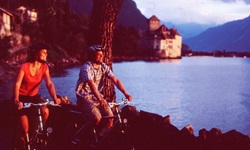 Ein Mann und eine Frau radeln am Ufer des Genfersees entlang. Im Bildhintergrund ist das Schloss Chillon zu sehen.
