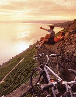 Ein Mann und eine Frau machen in einem Weinberg entlang der Rhone-Route Pause und schauen hinab auf den im Abendlicht liegenden Genfersee.