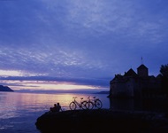 Ein Pärchen hat seine Fahrräder vor der Kulisse von Schloss Chillon am Ufer des Genfersees abgestellt und genießt das romantische Abendlicht.