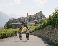 Ein Mann und eine Frau radeln auf dem Rhone-Radweg zwischen Weinreben hindurch und lassen den Ort Saillon hinter sich.