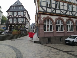 Eine Frau mit einem roten Poncho steht in einer Gasse einer alten Stadt entlang des Lahn-Radwegs und posiert lustig