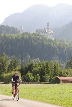 Ein Radfahrer fährt auf einem Radweg entlang, auf dem im Hintergrund das Schloss Neuschwanstein zu sehen ist.
