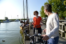 Ein Radlerpärchen steht auf einem Steg und unterhält sich, im Hintergrund liegen einige Boote vor Anker.