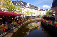 Ein Kanal der Saar schlängelt sich durch Saarburg - links und rechts vom Kanal Menschen, die in einem der Cafés unter den Sonnenschirmen sitzen
