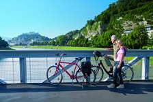 Zwei Radfahrer stehen auf einer Brücke über der Salzach und blicken nach Salzburg mit ihrer Festung