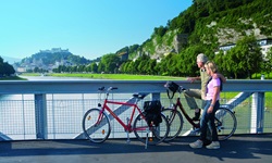 Zwei Radfahrer stehen auf einer Brücke über der Salzach und blicken nach Salzburg mit ihrer Festung
