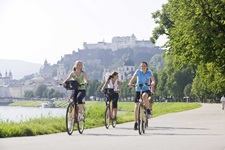 Drei junge Frauen radeln auf einem asphaltierten Radweg bei Salzburg. Im Hintergrund ist die Salzburger Skyline mit der Festung Hohensalzburg und dem Dom zu sehen.