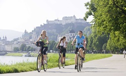 Drei junge Frauen radeln auf einem asphaltierten Radweg bei Salzburg. Im Hintergrund ist die Salzburger Skyline mit der Festung Hohensalzburg und dem Dom zu sehen.
