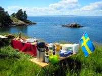 Ein appetitlich auf Holztischen angerichtetes und mit einer schwedischen Fahne dekoriertes Picknick wartet in einer idyllischen Bucht in Südschweden auf die hungrigen Radler.