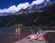 Eine Radlergruppe, die auf der Schweizer Seenroute unterwegs ist, macht an einem von Berggipfeln umgebenen See Pause. Ein Pärchen kommt gerade Hand in Hand aus dem Wasser, während ein anderes neben den abgestellten Rädern auf einem schmalen Kiesstrand sitzt.